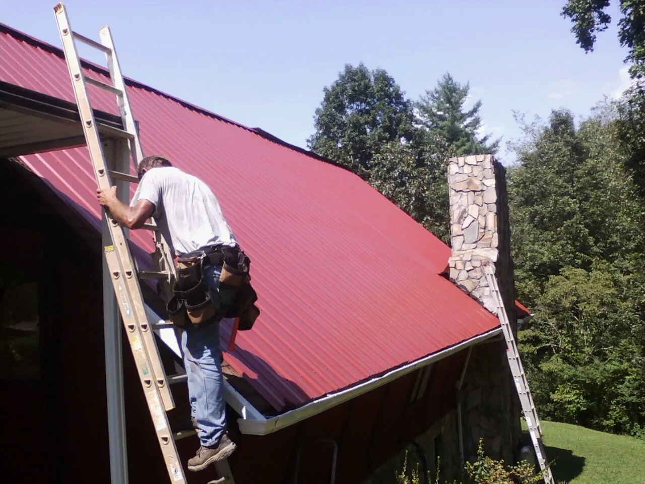 Roofer on a ladder beside a red metal roof and stone chimney — Soffit & Fascia Repair by Roofing Brothers in Shannock