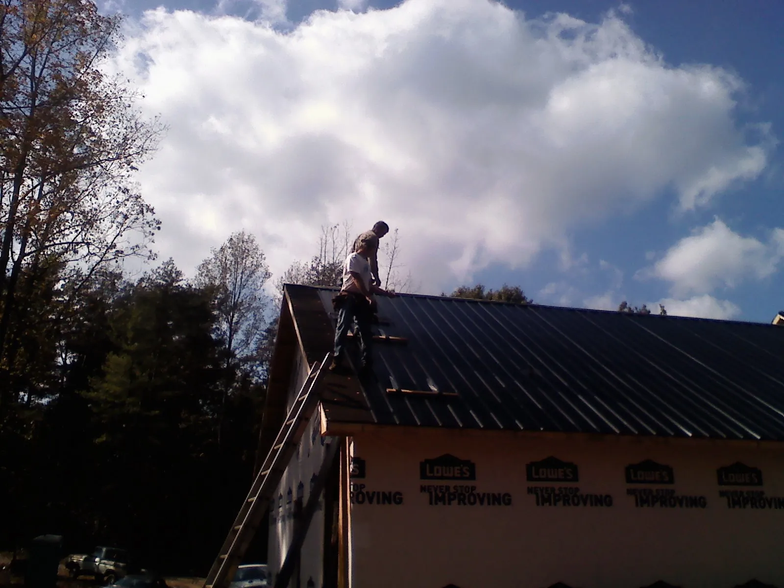 Roofer fastening dark metal panels high against a cloudy sky, from a Roof Inspection job in Shannock